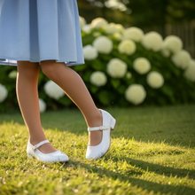 MOODBOARD kids white glossy Mary Jane shoes with buckle strap worn by a child in a garden setting.